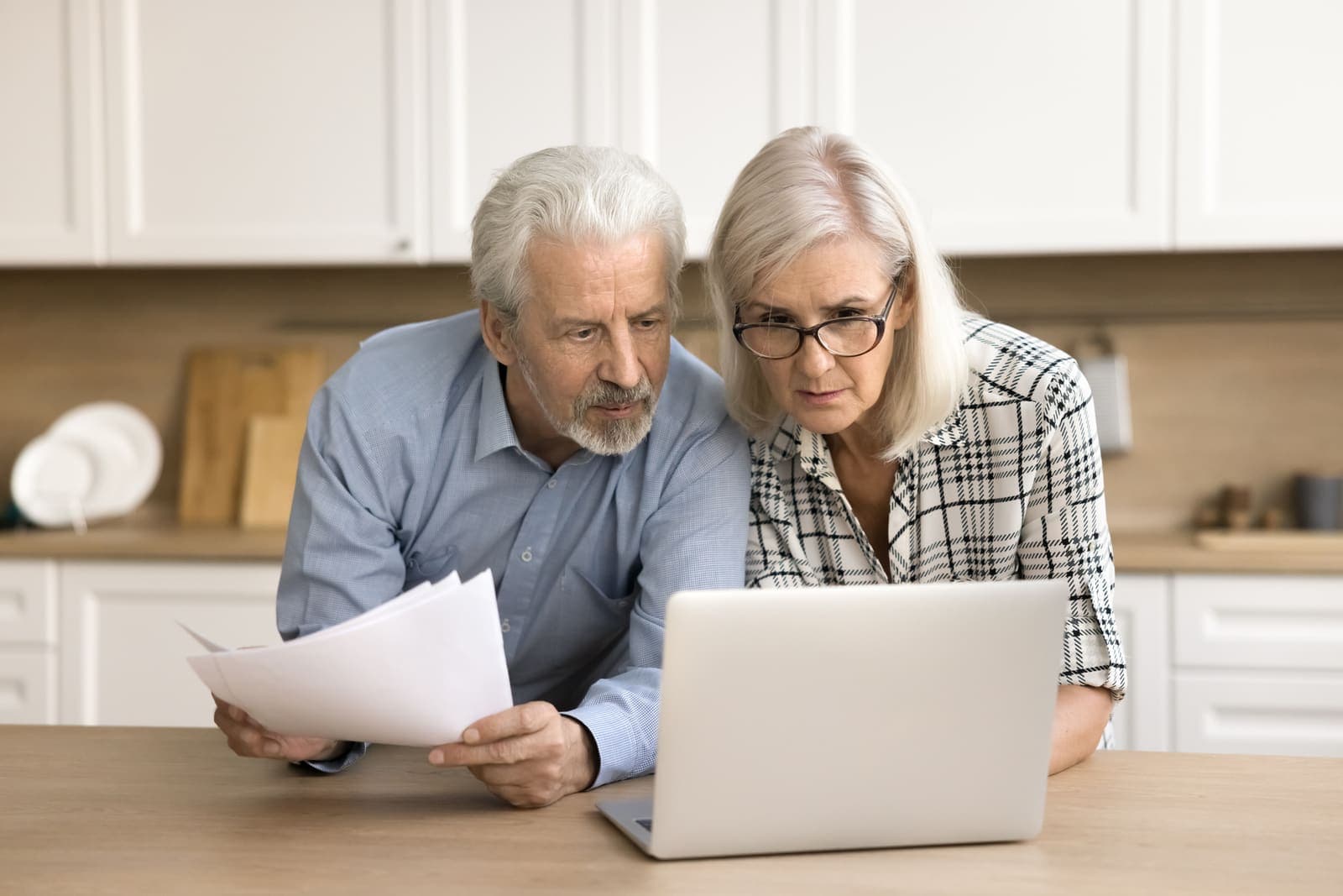 An older couple reviewing paperwork and a laptop at their kitchen table